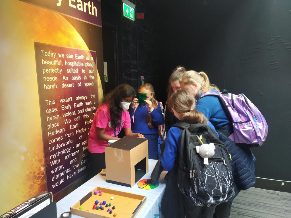 4 students are huddled around a science communicator. They are at a table exploring the concept of colour. A colour wheel, a number of coloured balls, and coloured light filters can all be seen.