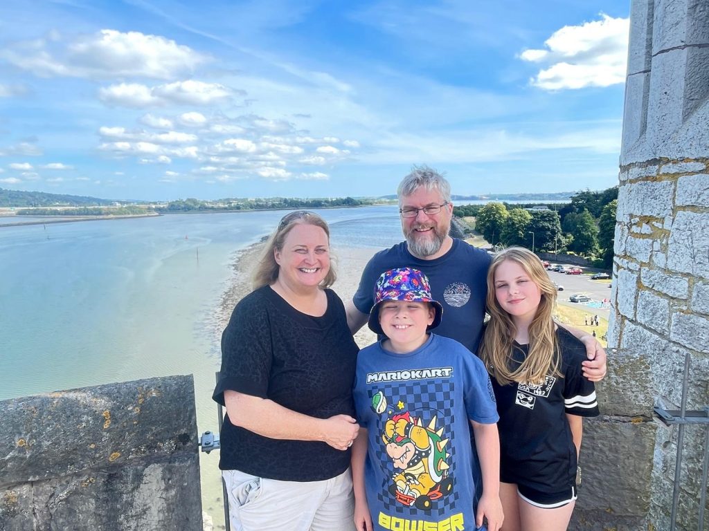 A family of four posing at the top of a castle tower overlooking Cork Harbour. The harbour dominates the photo with a beautiful blue sky reflected on the river behind the family.